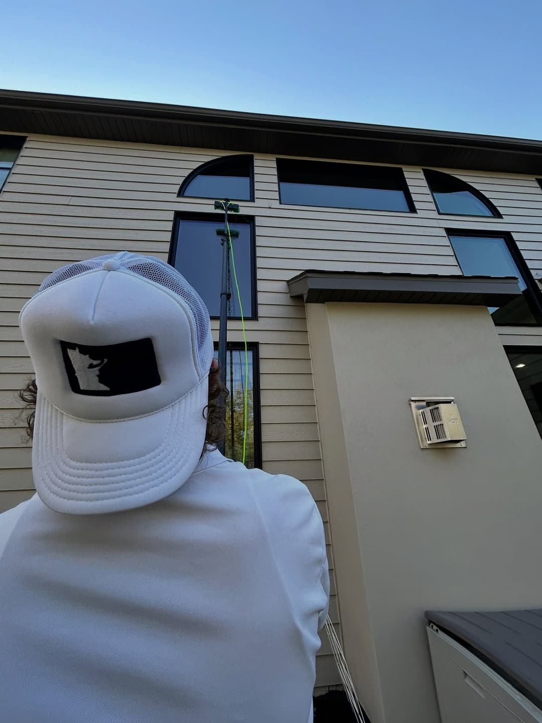 Person in a white hat cleaning a window on a modern house with a long-reaching tool.