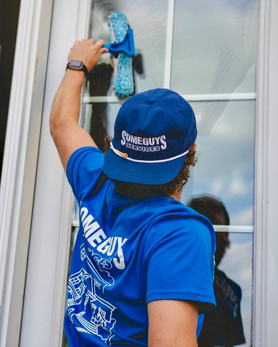 Person cleaning a window with a squeegee, wearing a blue "Some Guys Services" cap and shirt.