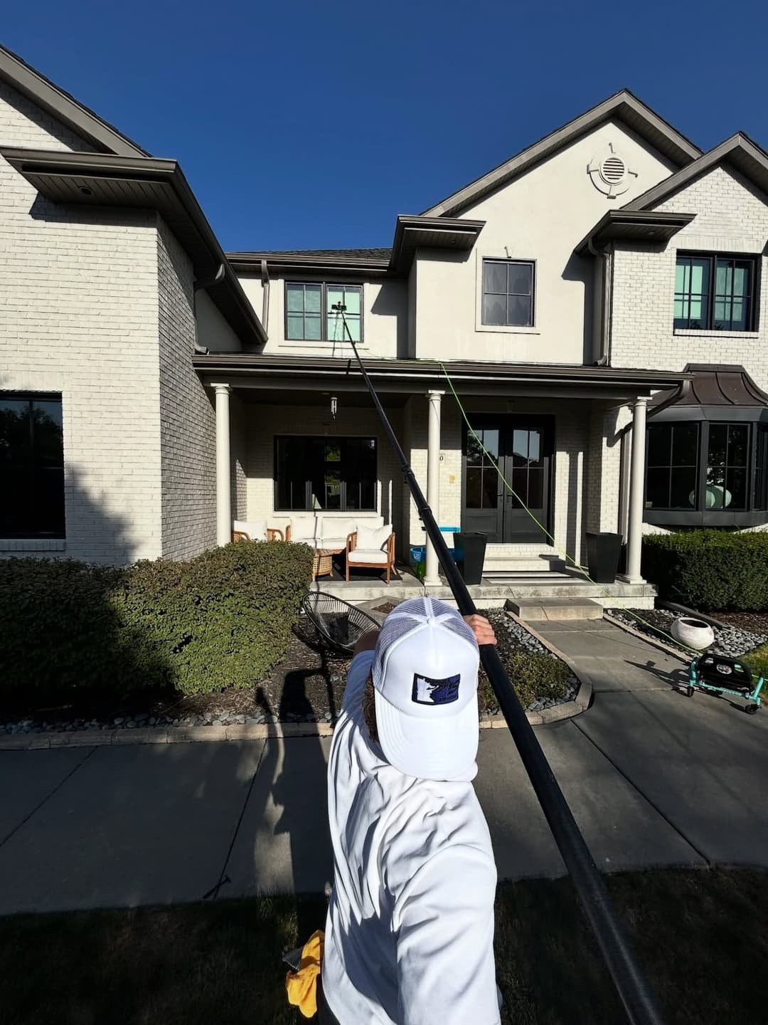 Person using a pole to clean a window on a two-story house under clear blue sky.