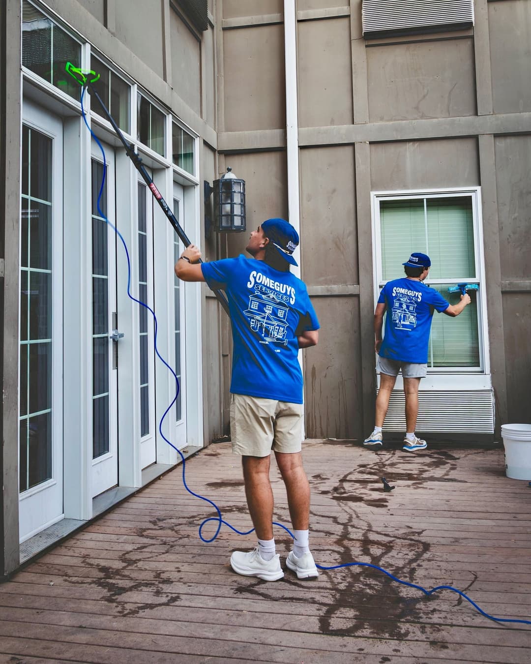 Men in blue shirts cleaning windows with a squeegee and extension poles on a wood deck.