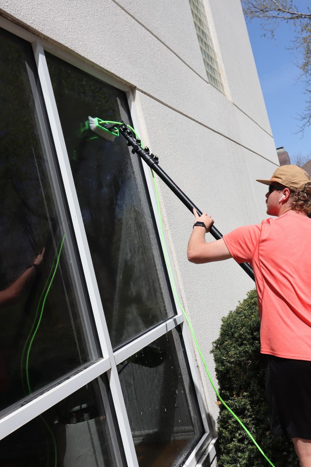 Man cleaning windows with a long reach tool on a sunny day near a building.