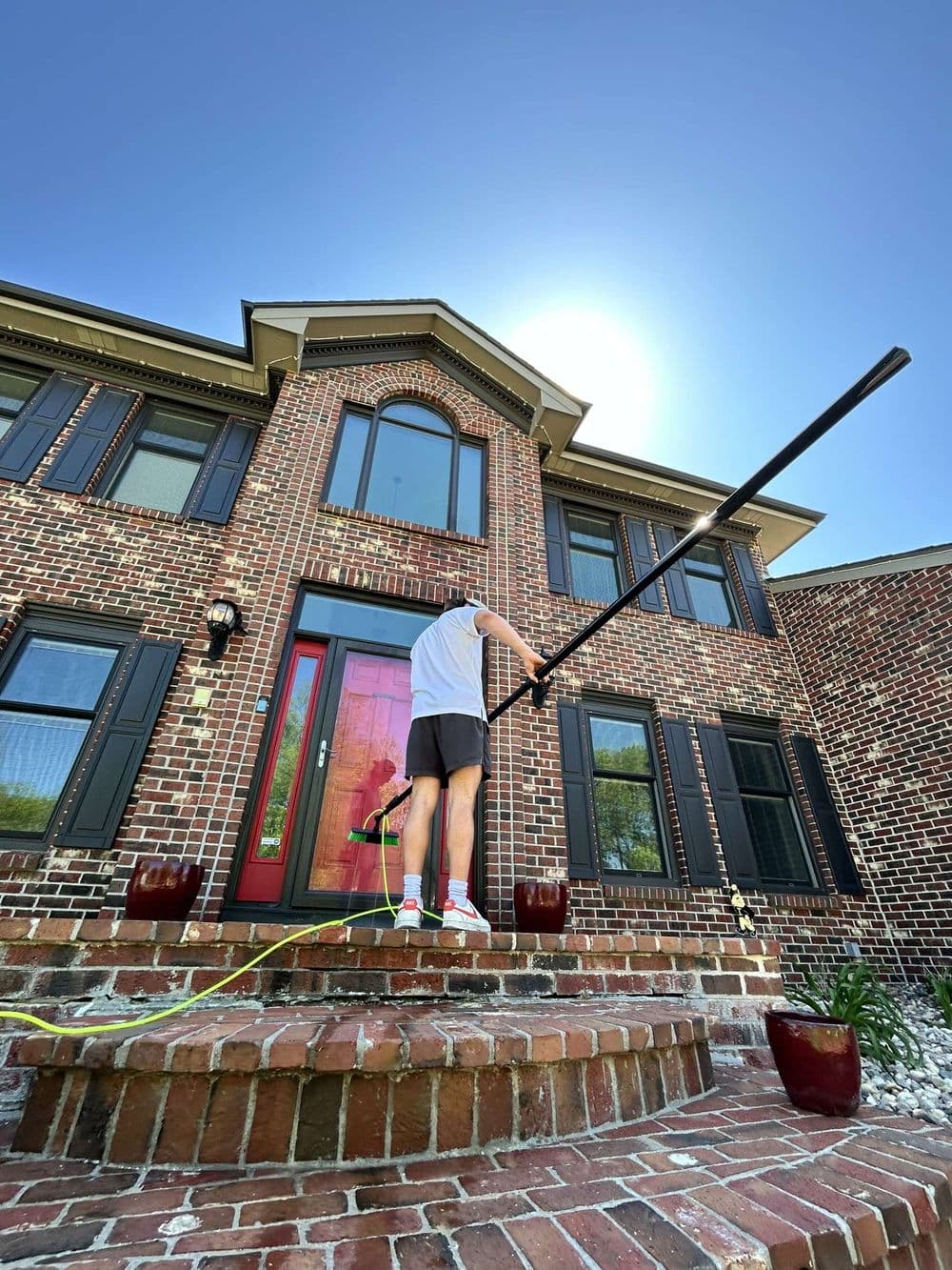 Person cleaning a brick house exterior with a long pole beside a red front door.