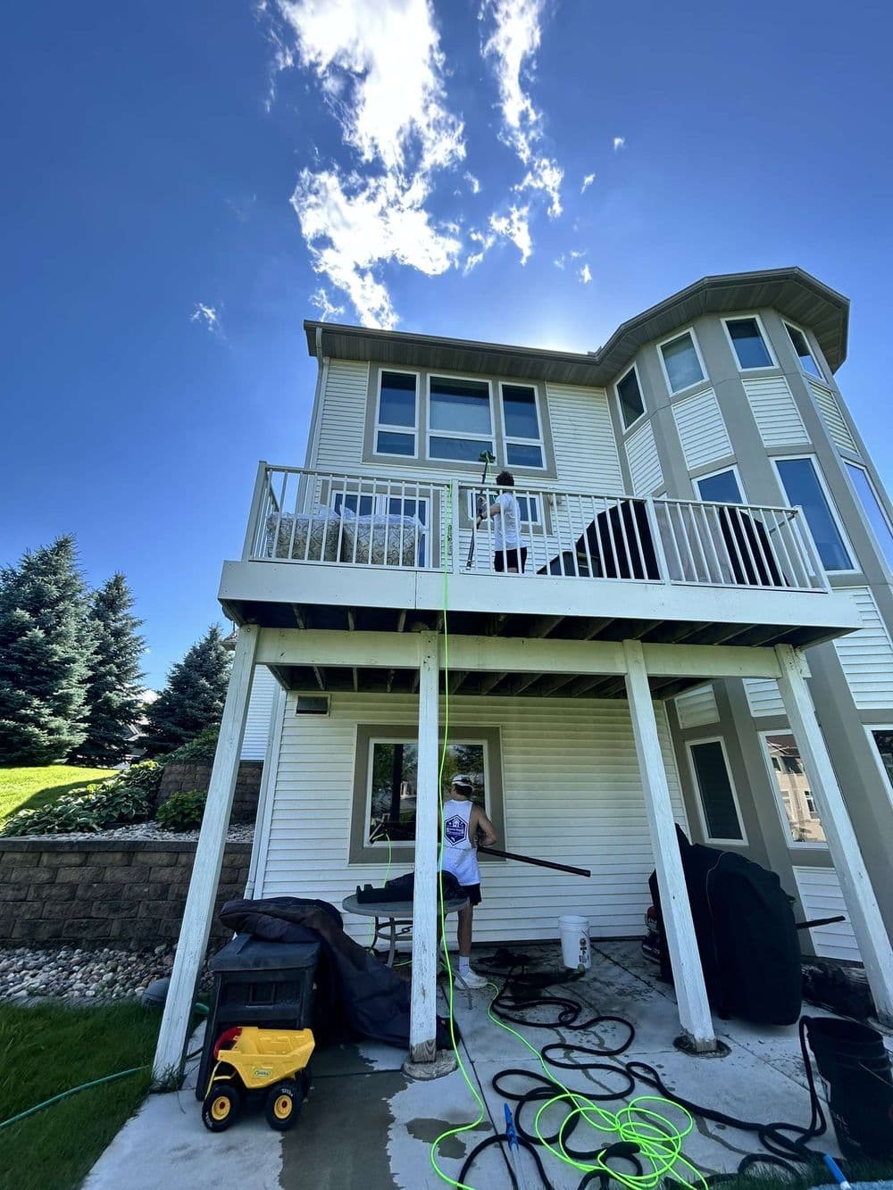 Person cleaning the exterior of a two-story house with a pressure washer under a blue sky.