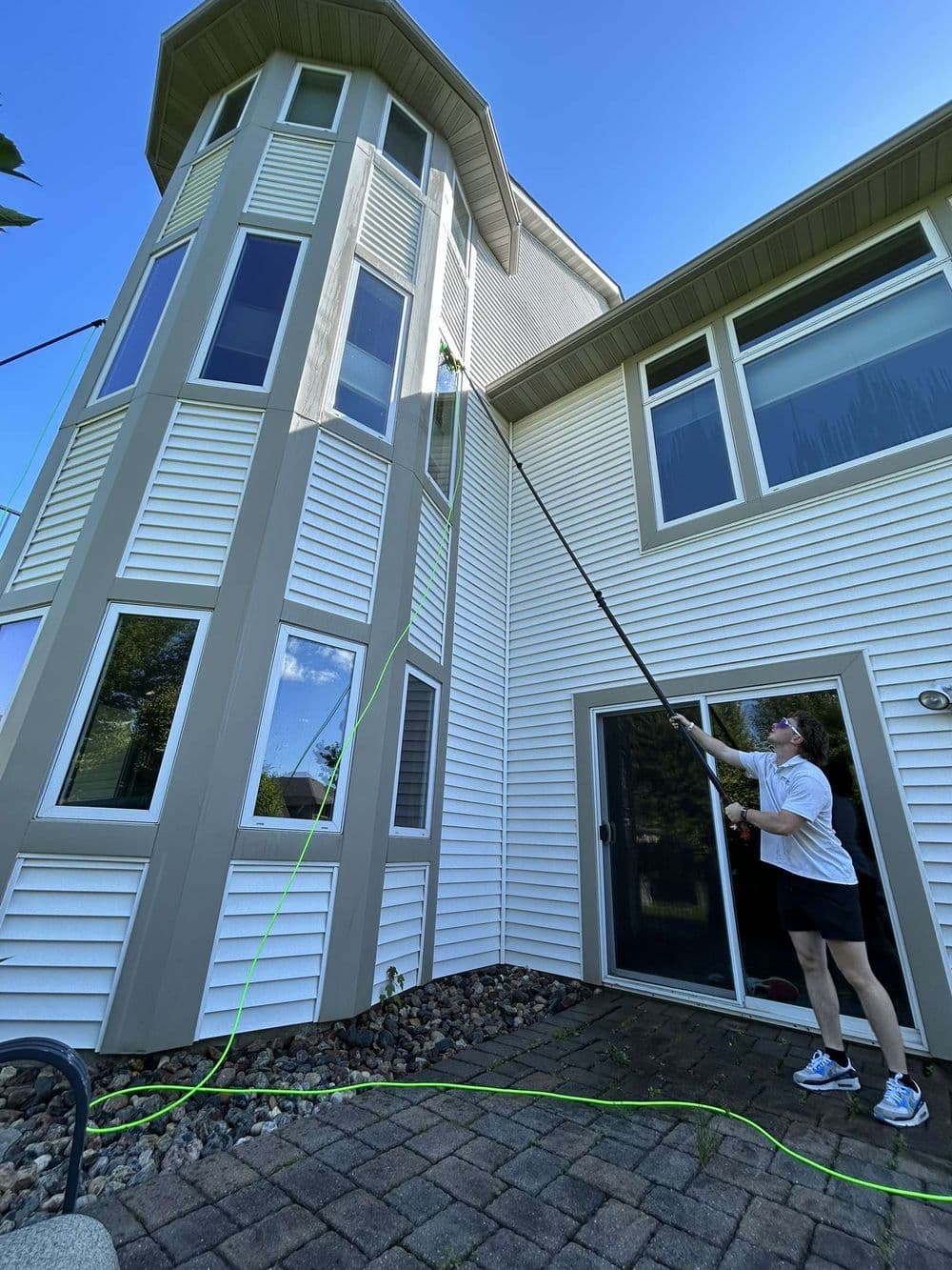 Person cleaning a tall house exterior with a long pole and green hose on a clear day.