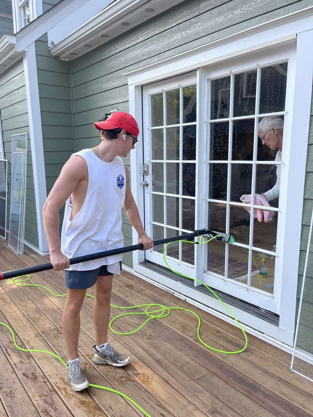 Young man washing windows with a garden hose, while an older person watches from inside a house.