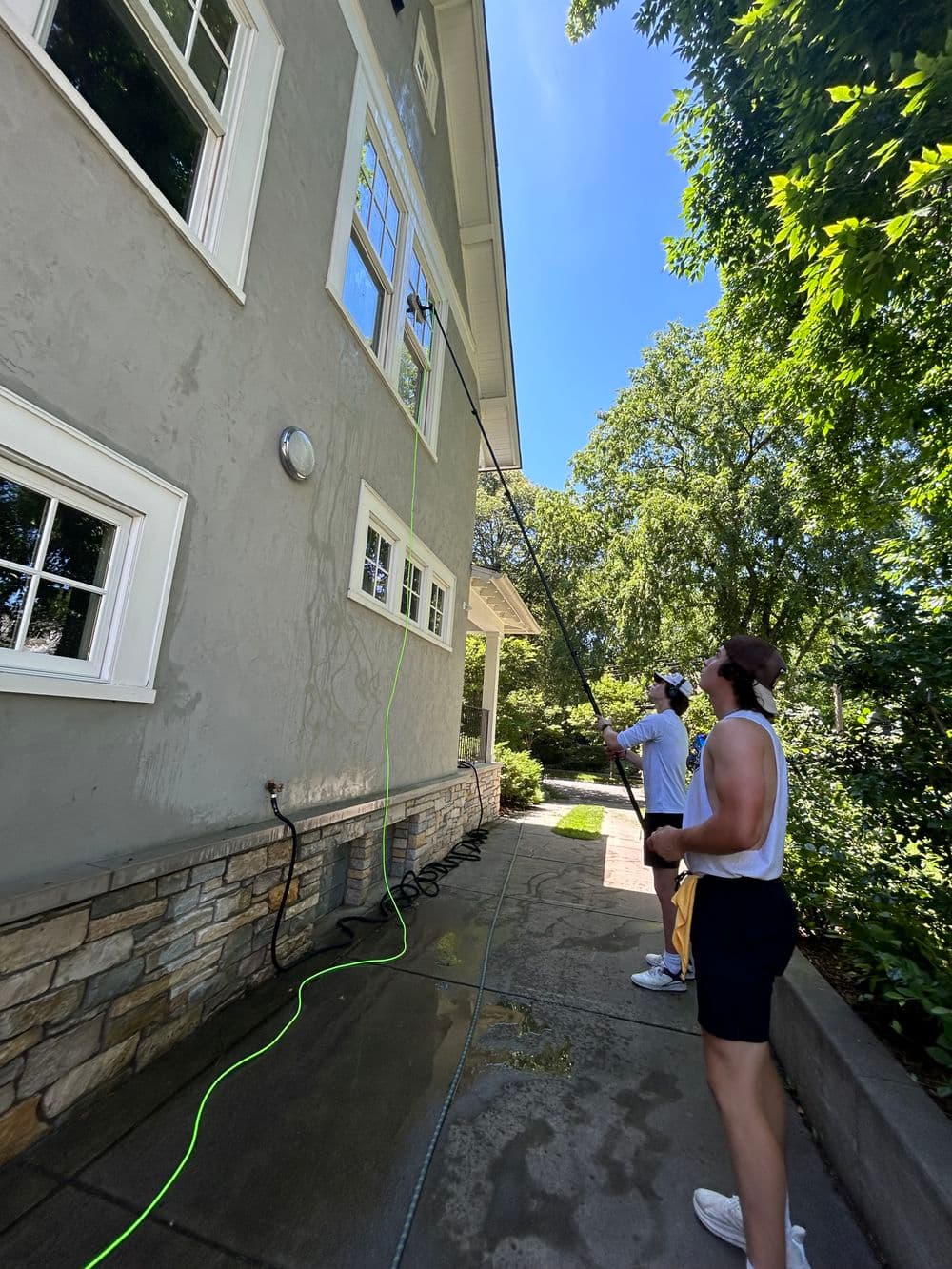 Two men using a long pole to wash windows on a house under sunny skies.