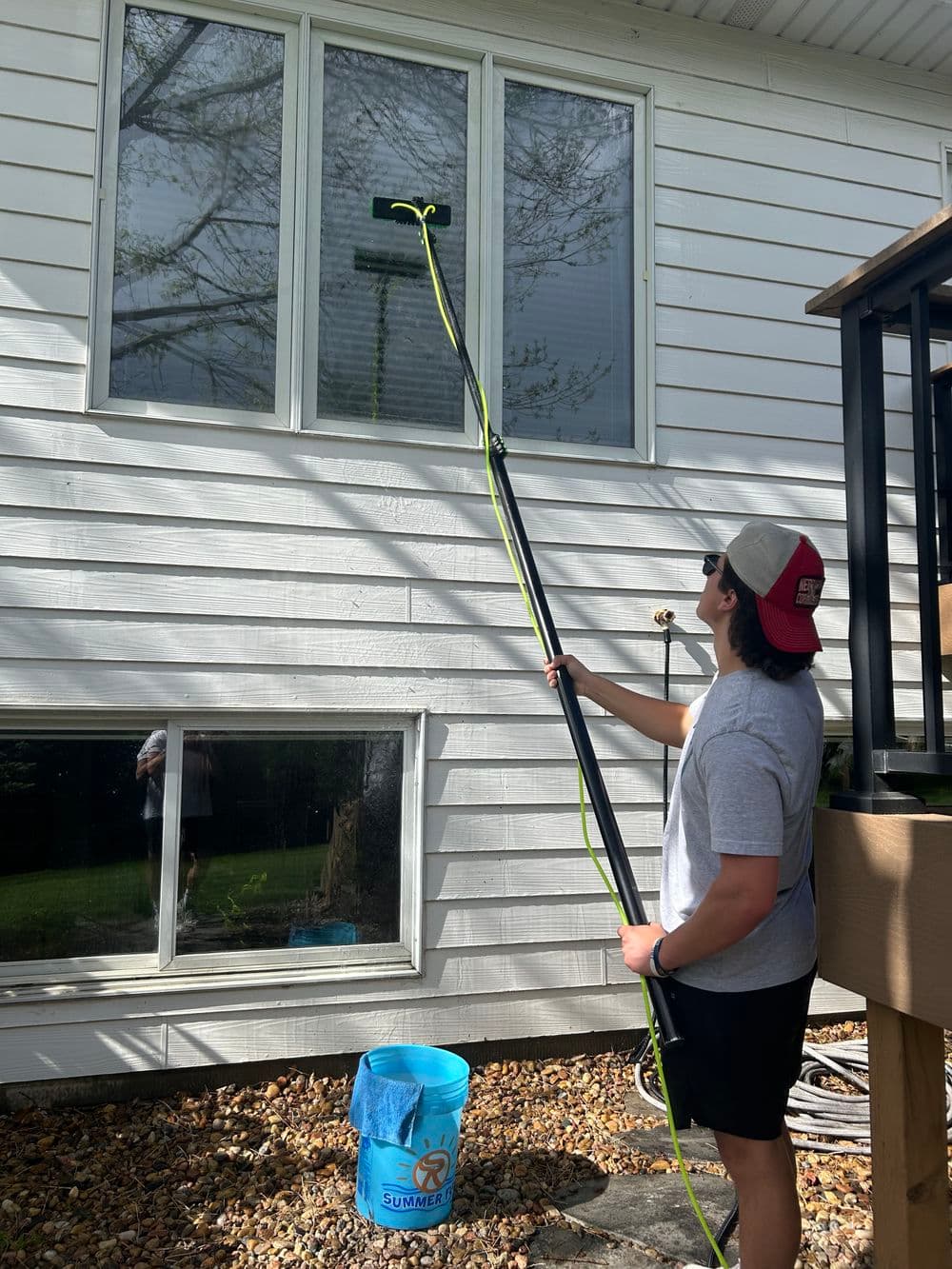 Person using a long pole to clean windows on a house exterior on a sunny day.