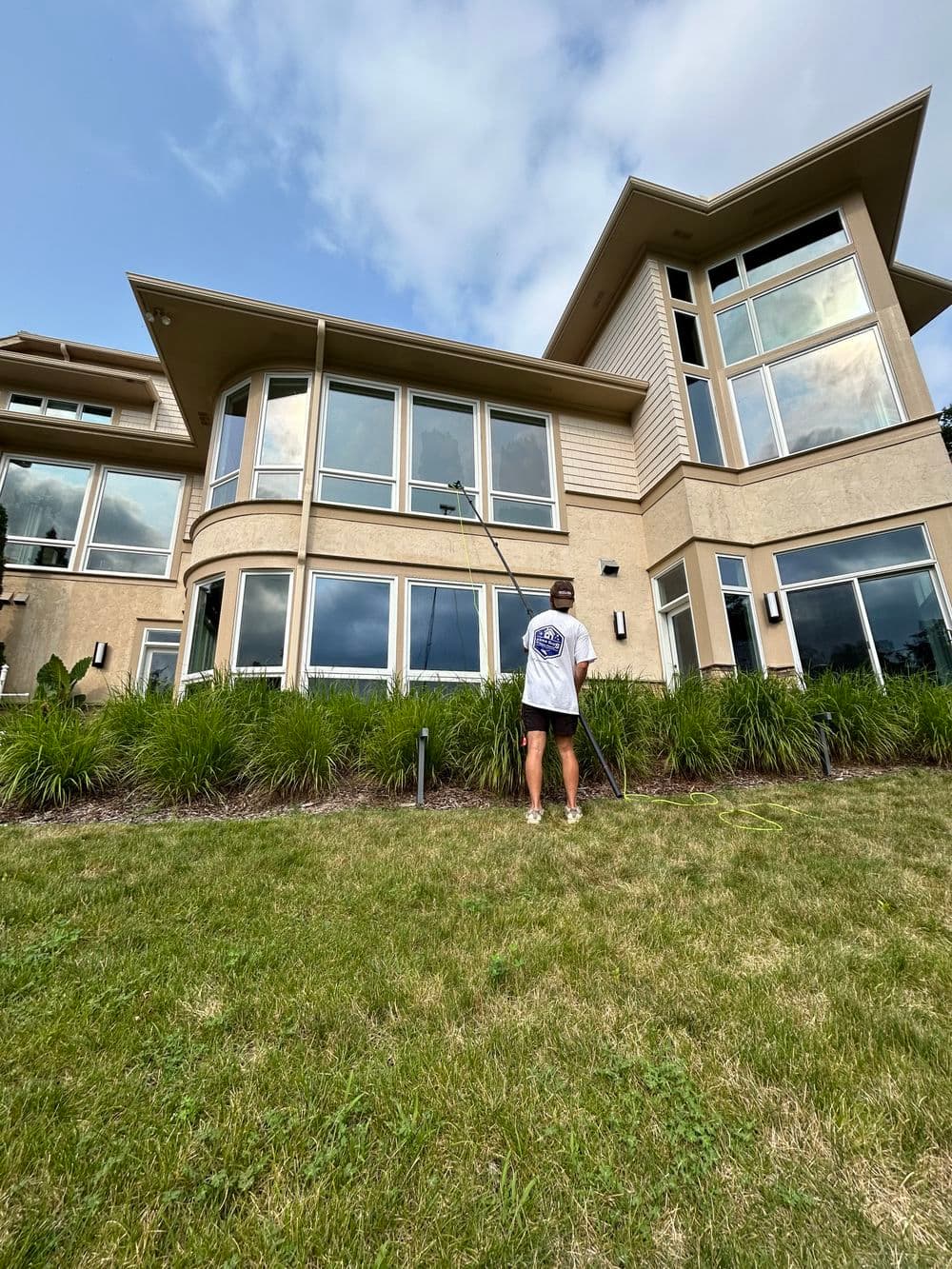 Person cleaning large windows on a modern house with multiple glass panels and greenery.