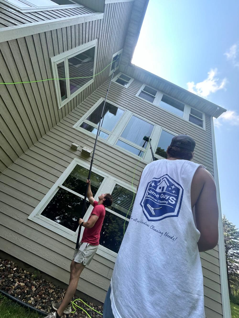Two workers using a long pole to clean windows on a two-story home exterior, sunny day.