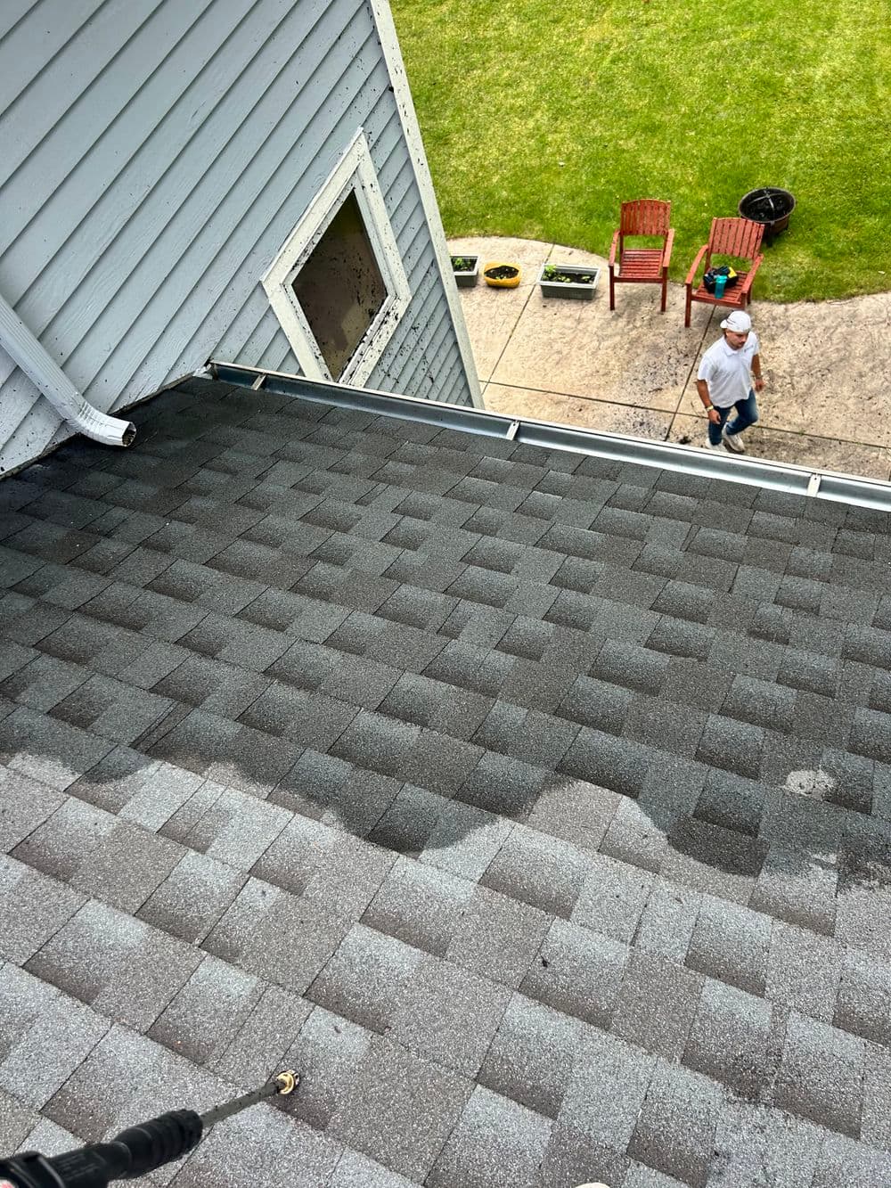 Person cleaning a residential roof with pressure washer, green lawn and chairs in background.